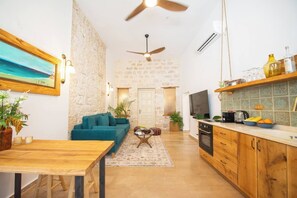 Ein zeitgemäßer Wohnraum mit Steinwand, einem Salbeigrünen Sofa, einem Holzesstisch und einer Küchenzeile mit Fernseher und diversen Küchengeräten.