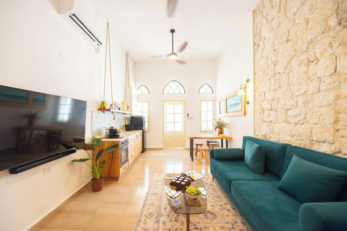 Una sala de estar con un sofá azul verdoso, un televisor de pantalla plana, una cocina con microondas y una pared de piedra.