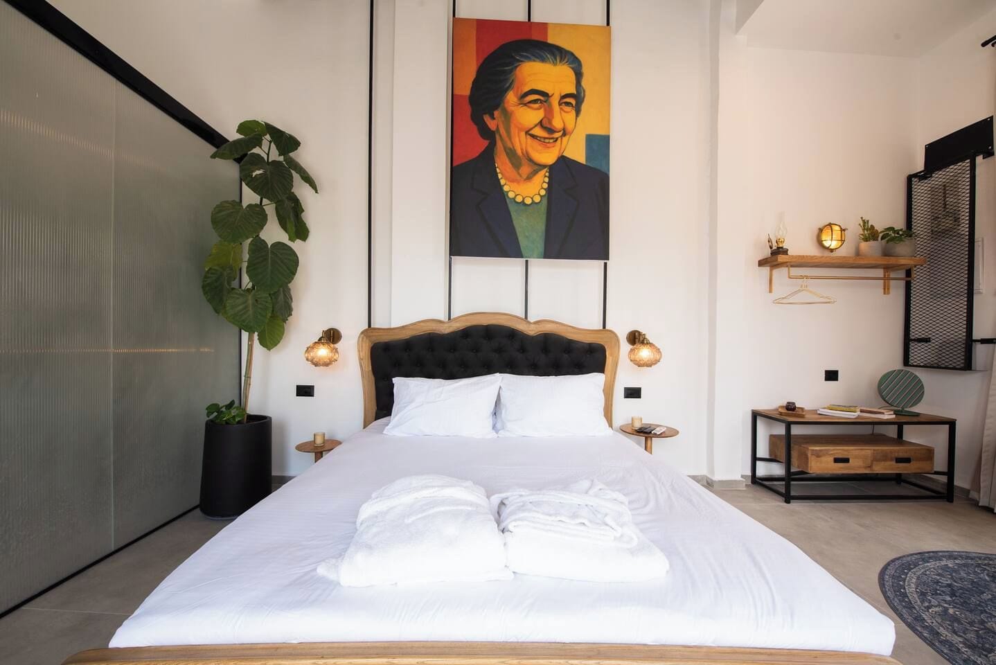 Un lit bien fait, avec du linge de lit et des serviettes blanches, une grande plante en pot, un tableau accroché au mur et une petite table de chevet avec une lampe.