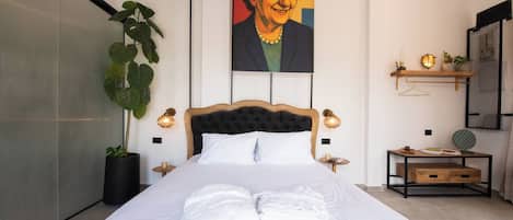 Un lit bien fait, avec du linge de lit et des serviettes blanches, une grande plante en pot, un tableau accroché au mur et une petite table de chevet avec une lampe.