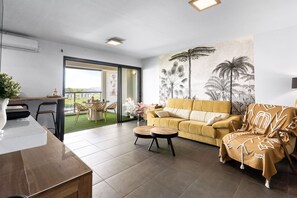 Ein modernes Wohnzimmer mit einem gelben Sofa, einem Couchtisch und einem Essbereich mit Tisch und Stühlen. Ein großes Fenster bietet einen Blick ins Freie.