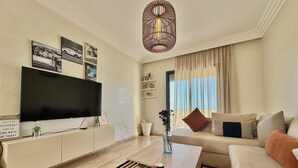 A modern living room with a flat-screen TV, a wooden coffee table, a sofa, and a large window with curtains. - 2 Bedroom Apartment Sea View 🌊 + Free Parking | Rabat (Rabat)