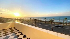 A balcony with a checkered floor overlooking a coastal road with palm trees and a sunset. - 2 Bedroom Apartment Sea View 🌊 + Free Parking | Rabat (Rabat)