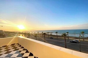 A balcony with a checkered floor overlooking a coastal road with palm trees and a sunset. - 2 Bedroom Apartment Sea View 🌊 + Free Parking | Rabat (Rabat)