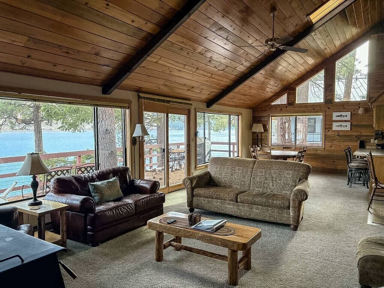 Ein gemütliches Blockhauss-Interieur mit Holzbalkendecke, zwei Sofas, einem Couchtisch und großen Fenstern mit Blick auf ein Gewässer.