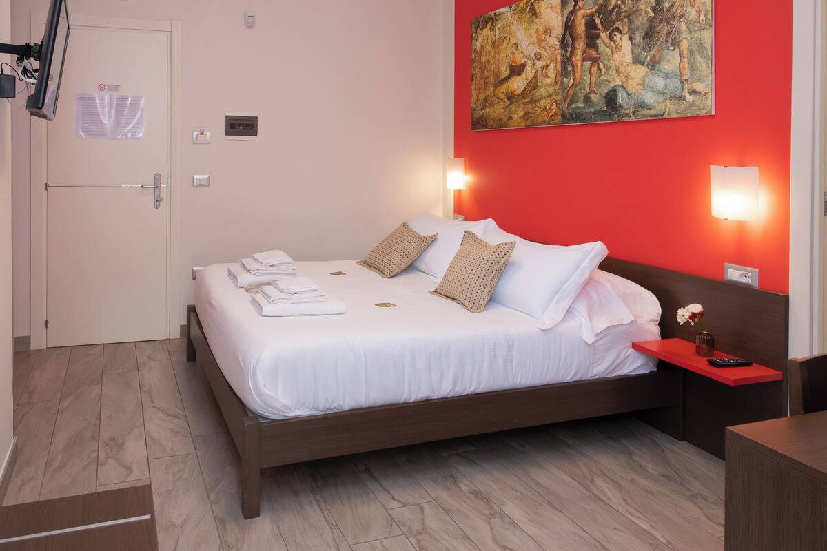 A hotel room with a bed, a bedside table, a wall-mounted painting, and a door with a sign.