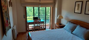 Ein Schlafzimmer mit einem Bett, einem Schreibtisch mit Laptop, einem Sessel, einem Nachttisch und einem Fenster mit Blick ins Grüne.