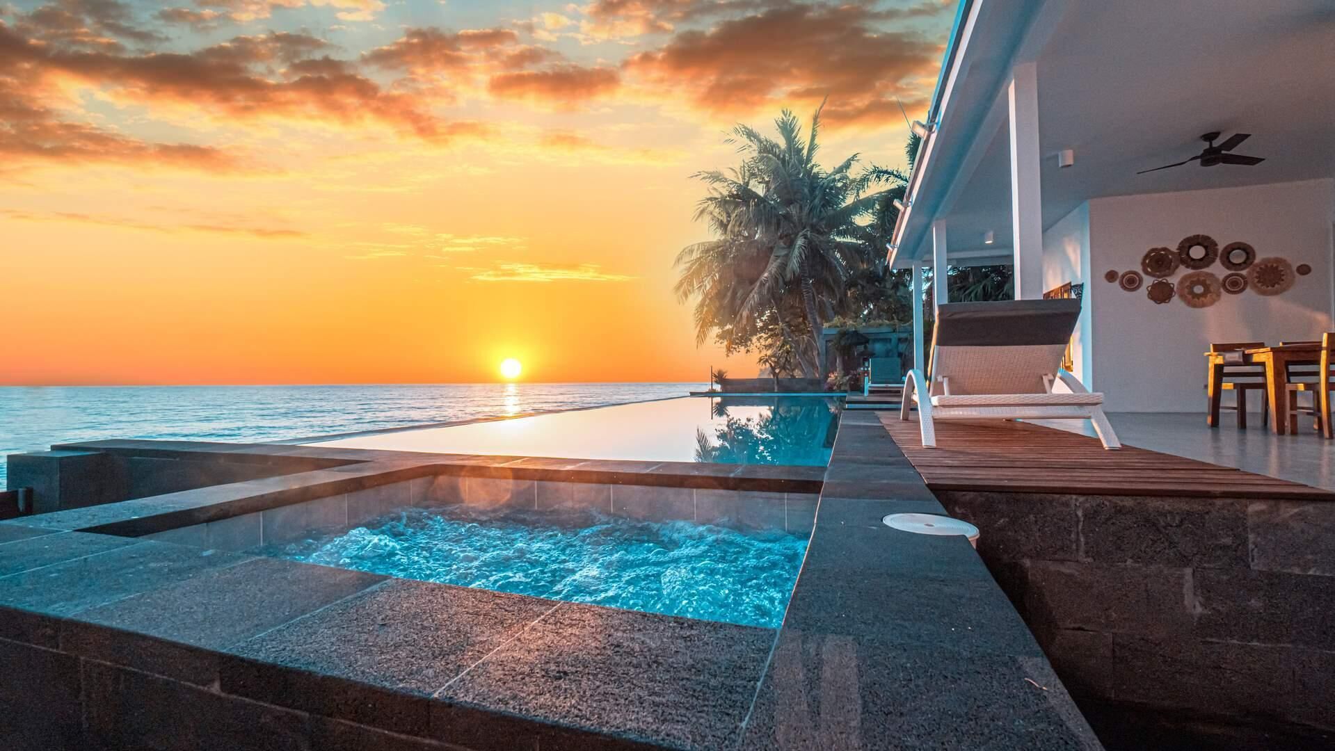 A pool area with a sunset view, lounge chairs, and a palm tree.
