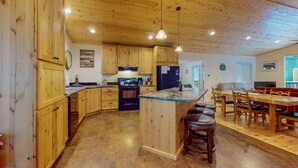 A kitchen with wooden cabinets, a central island with stools, a dining area with a table and chairs, and a visible refrigerator. - Perfect Getaway for Up to 8 Guests | Glacier Lake (Joseph)