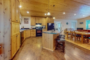 Eine Küche mit Holzausstattung, einer zentralen Kochinsel mit Hockern, einem Essbereich mit Tisch und Stühlen sowie einem sichtbaren Kühlschrank.