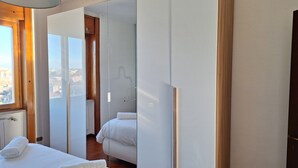 Ein modernes Hotelzimmer mit einem großen Bett, einem begehbaren Kleiderschrank und Blick auf die Stadt durch ein großes Fenster.
