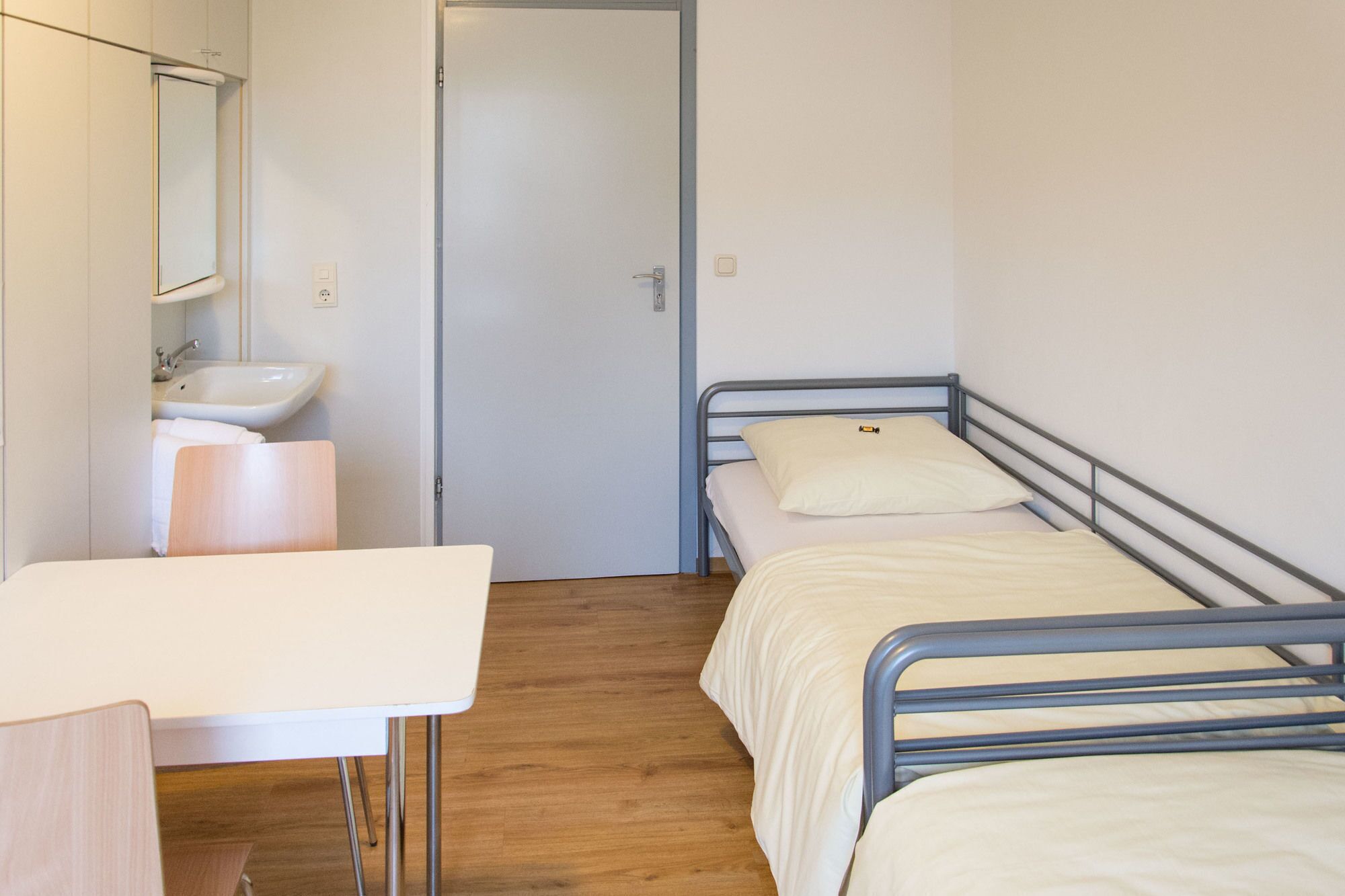 Une petite chambre avec un lit simple, un plancher en bois, une table de chevet, un lavabo blanc et un miroir.