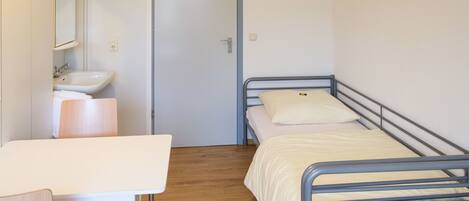 Une petite chambre avec un lit simple, un plancher en bois, une table de chevet, un lavabo blanc et un miroir.