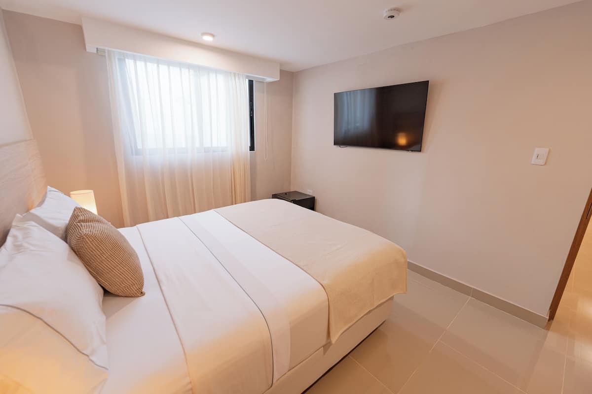 A hotel room with a bed, a bedside table, a flat-screen TV mounted on the wall, and a window with sheer curtains.