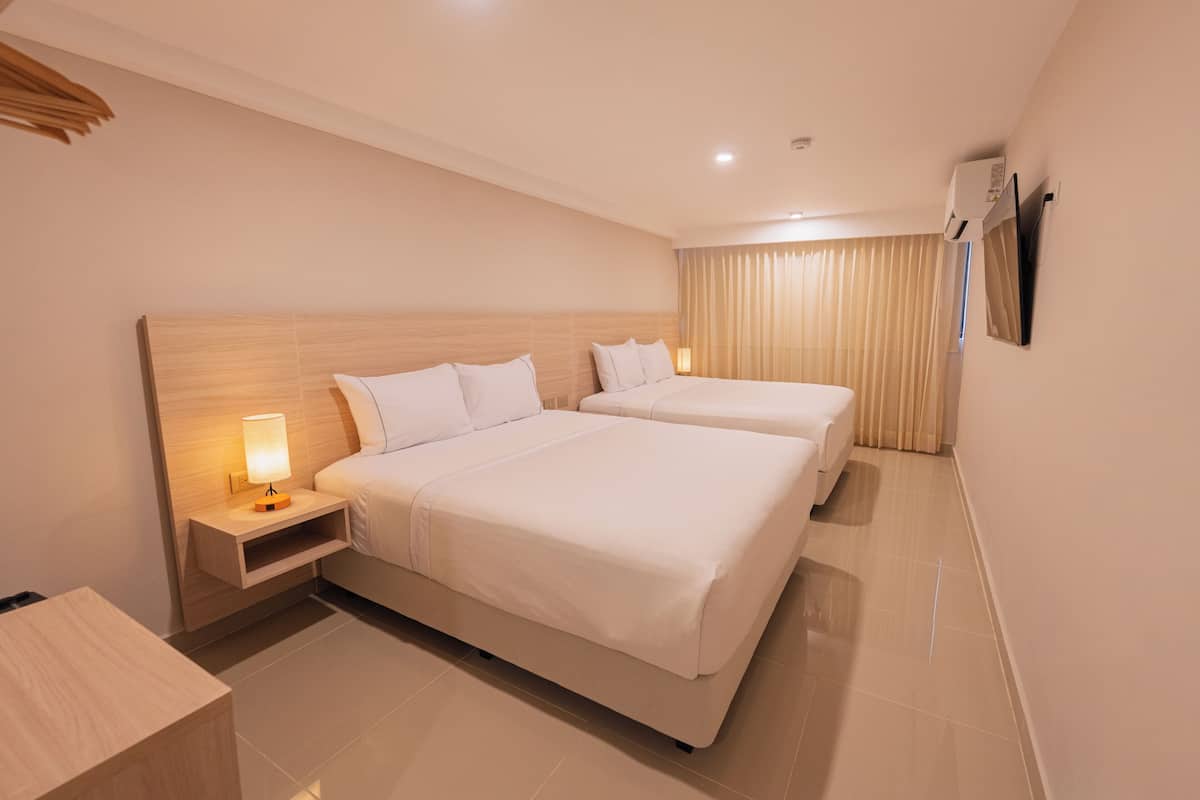 A hotel room with two beds, a bedside table with a lamp, a flat-screen TV mounted on the wall, and a window with curtains.