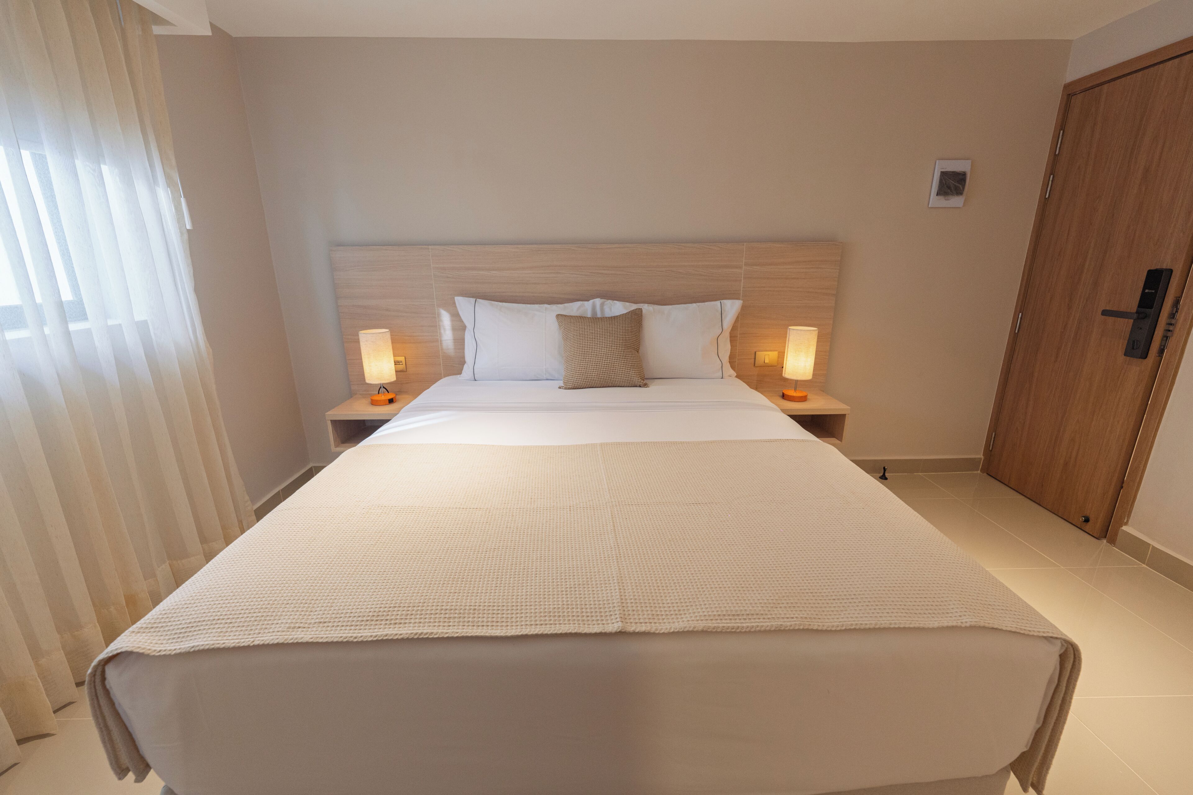 A hotel room with a large bed, two bedside lamps, a wooden headboard, and a wooden door.
