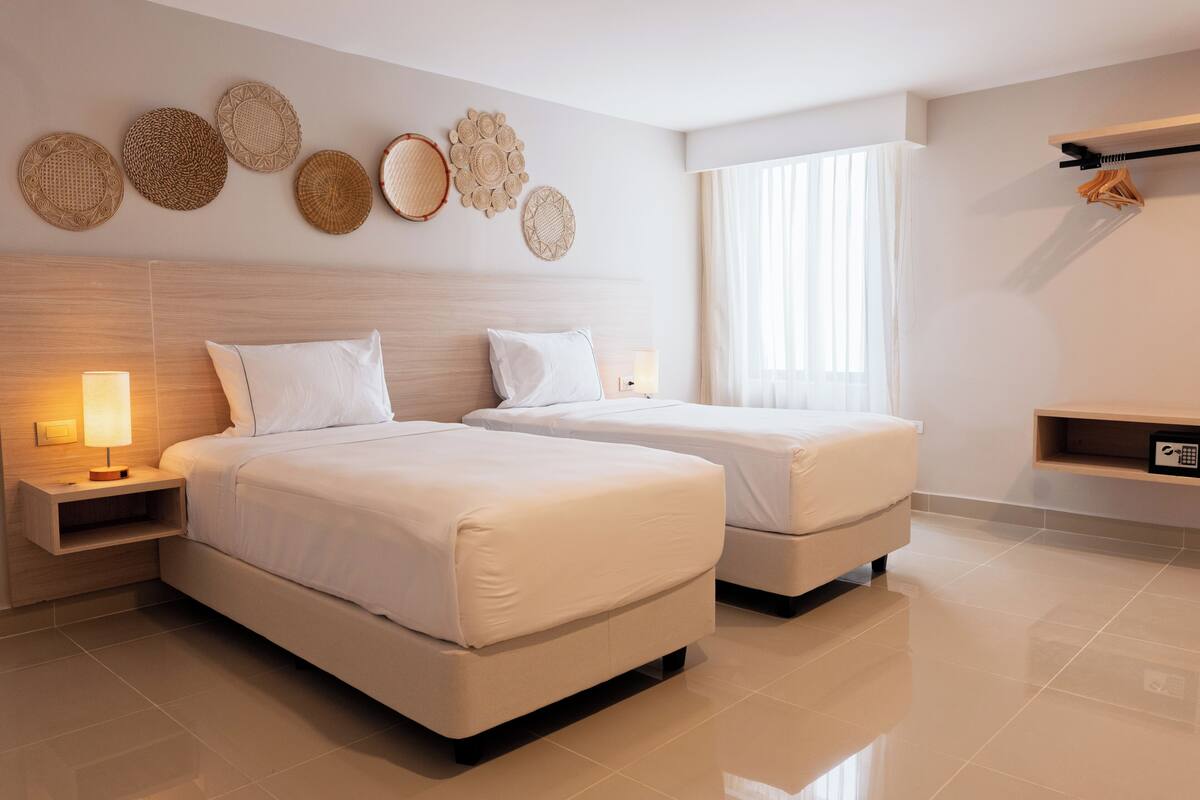 A hotel room with two beds, a bedside table with a lamp, a wall-mounted shelf, and a large window with curtains.
