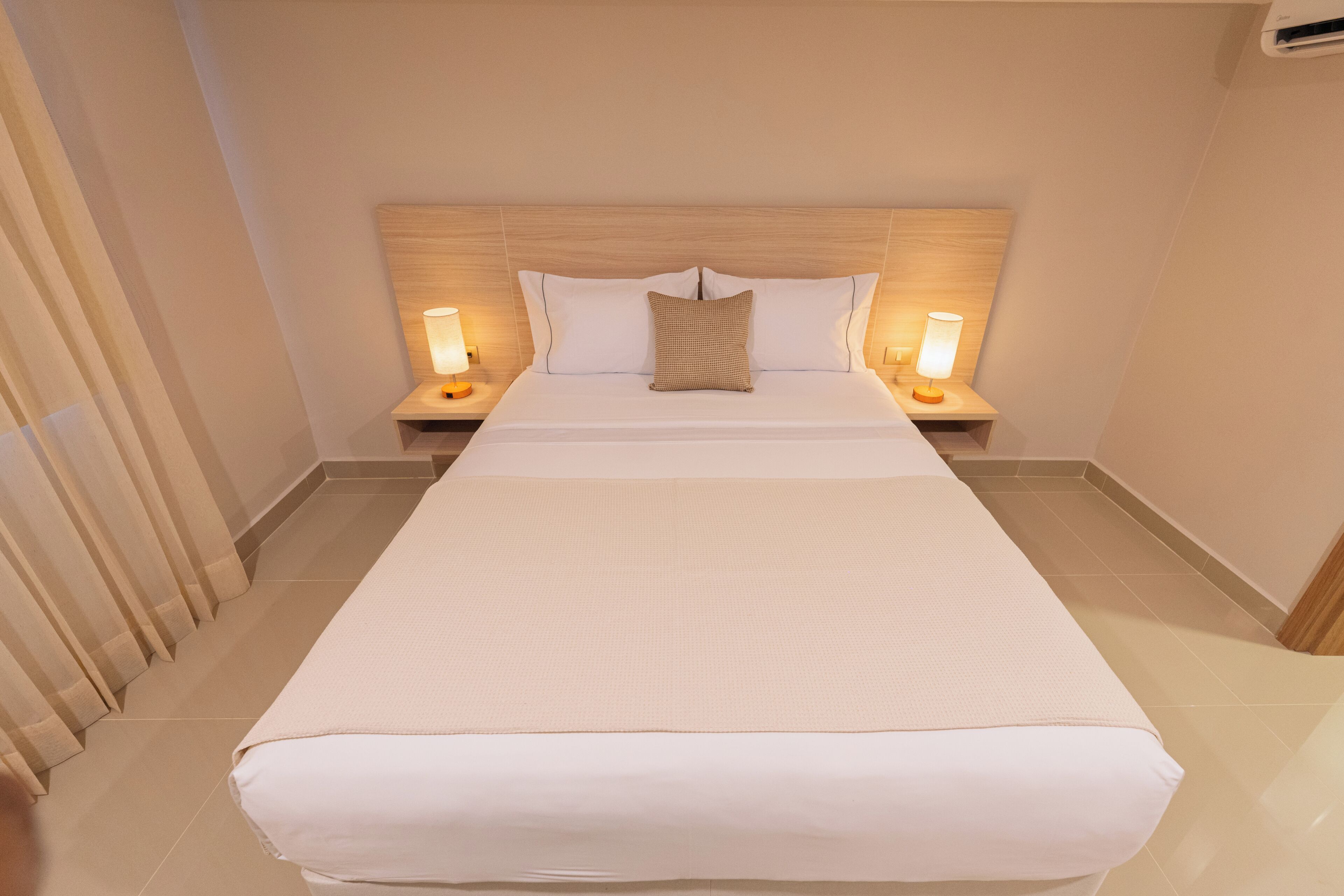 A hotel room with a large bed, two bedside tables with lamps, a wooden headboard, and a tiled floor.