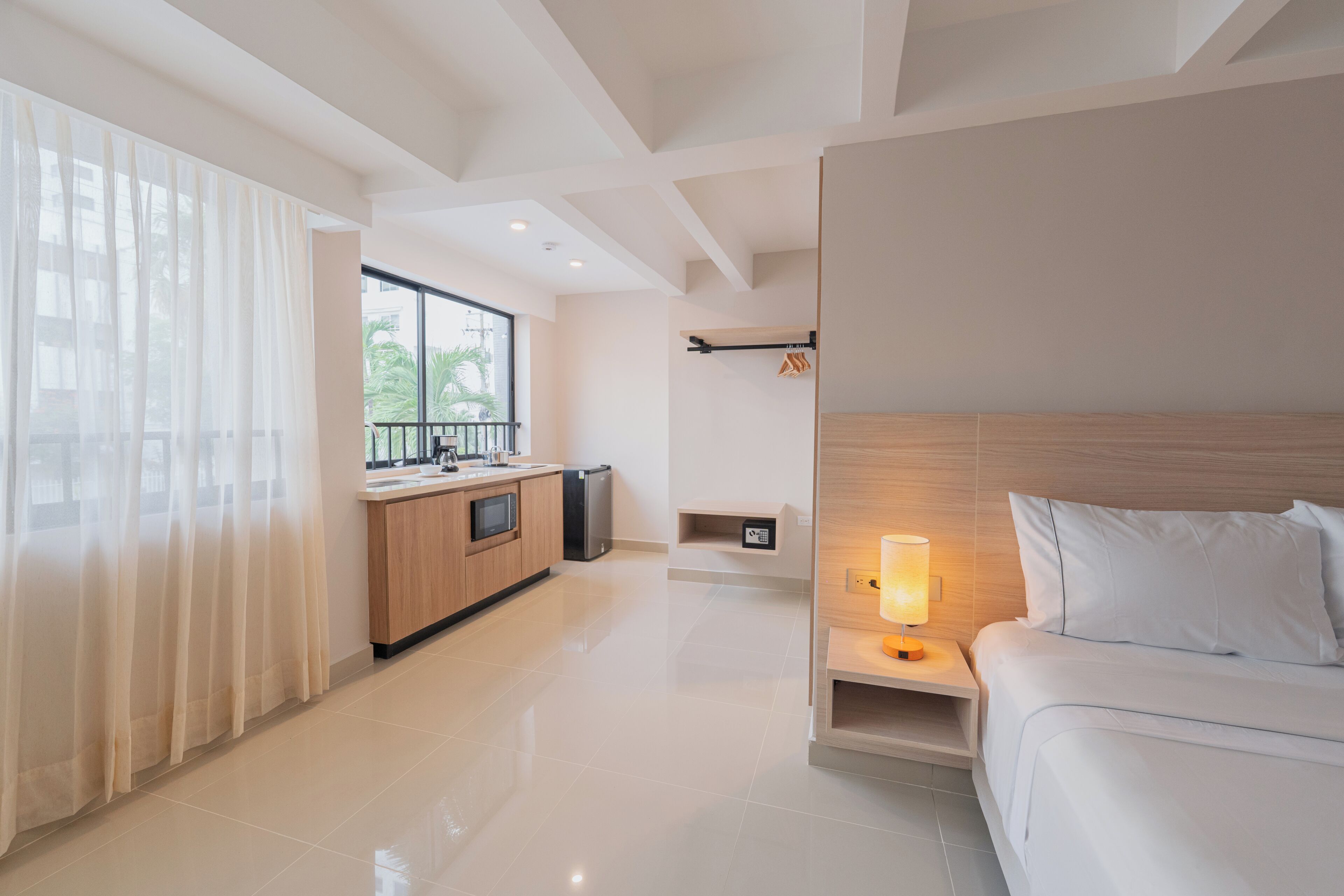 A modern hotel room with a large bed, a bedside table with a lamp, a wooden headboard, a kitchen area with a sink and cabinets, and a view of the outdoors through large windows.