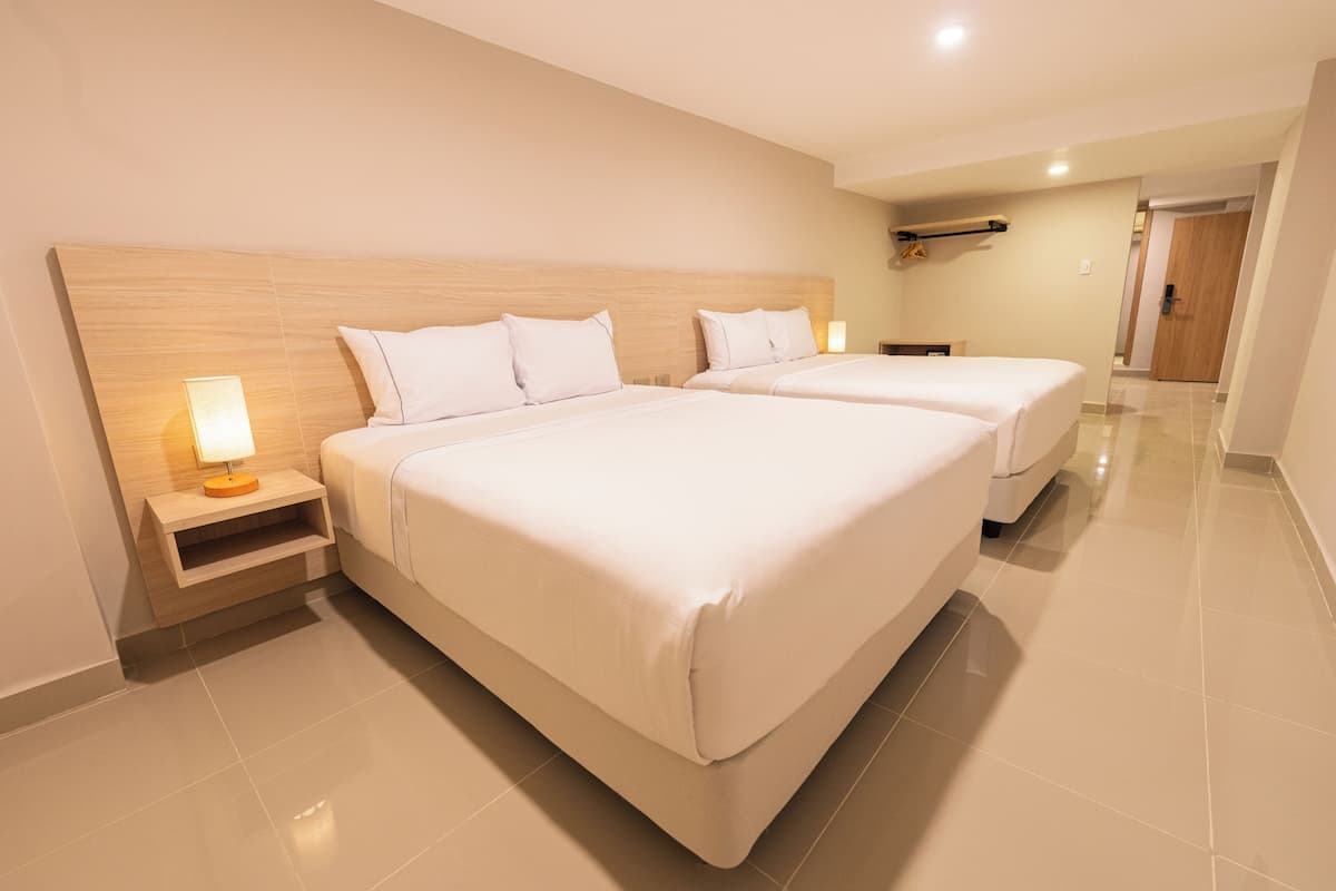 A hotel room with two beds, a nightstand with a lamp, a wooden headboard, and a bathroom door in the background.