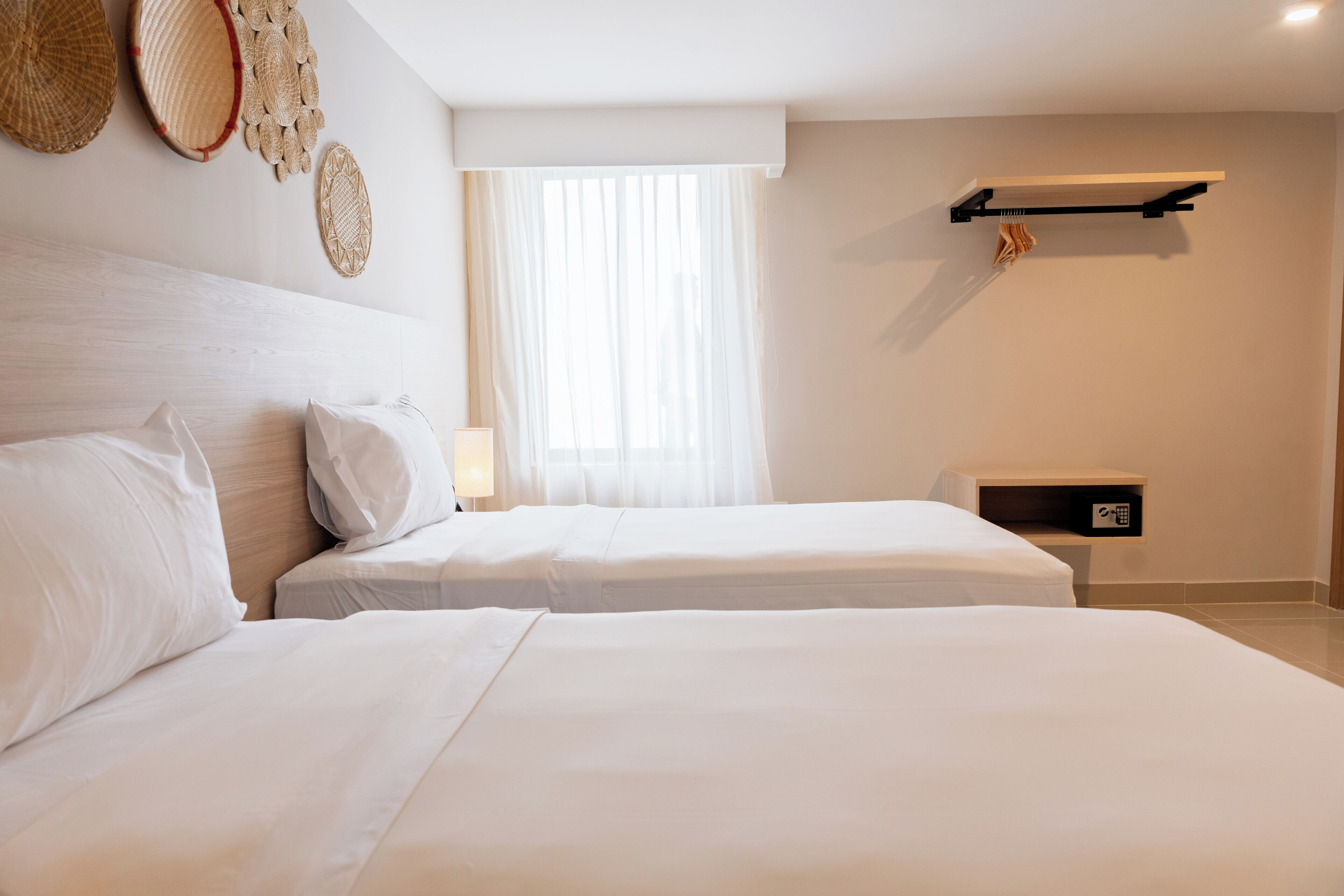Habitación de hotel con dos camas, una ventana con cortinas translúcidas, un estante en la pared y una mesita de noche.