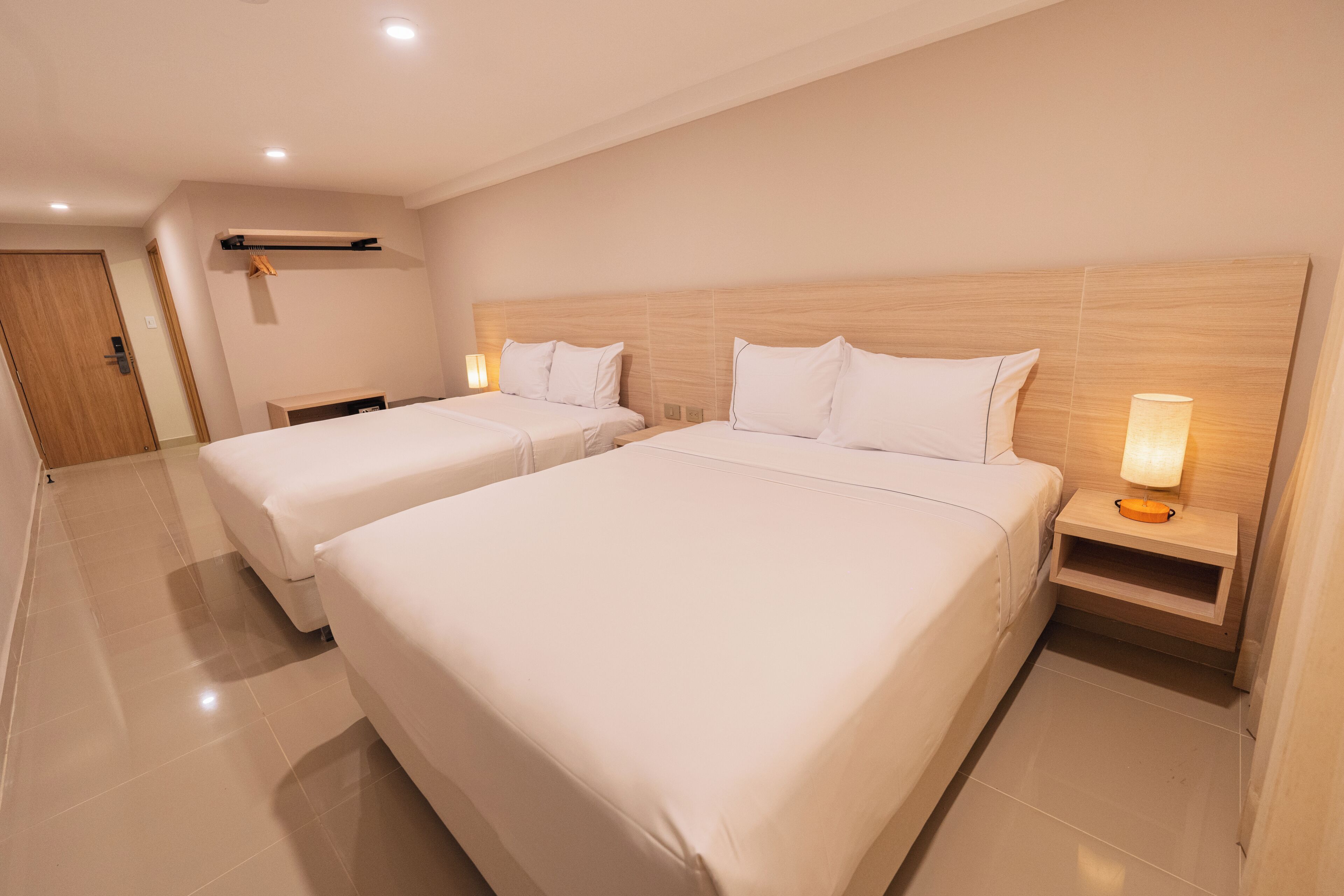 Une chambre d’hôtel avec deux lits, une tête de lit en bois, des lampes de chevet, un banc en bois et une porte.