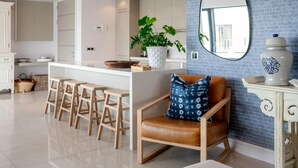 A modern kitchen with a central island, bar stools, a seating area with a cushioned chair, and a decorative mirror on the wall. - Le Paradis Penthouse Apartment (Hermanus)