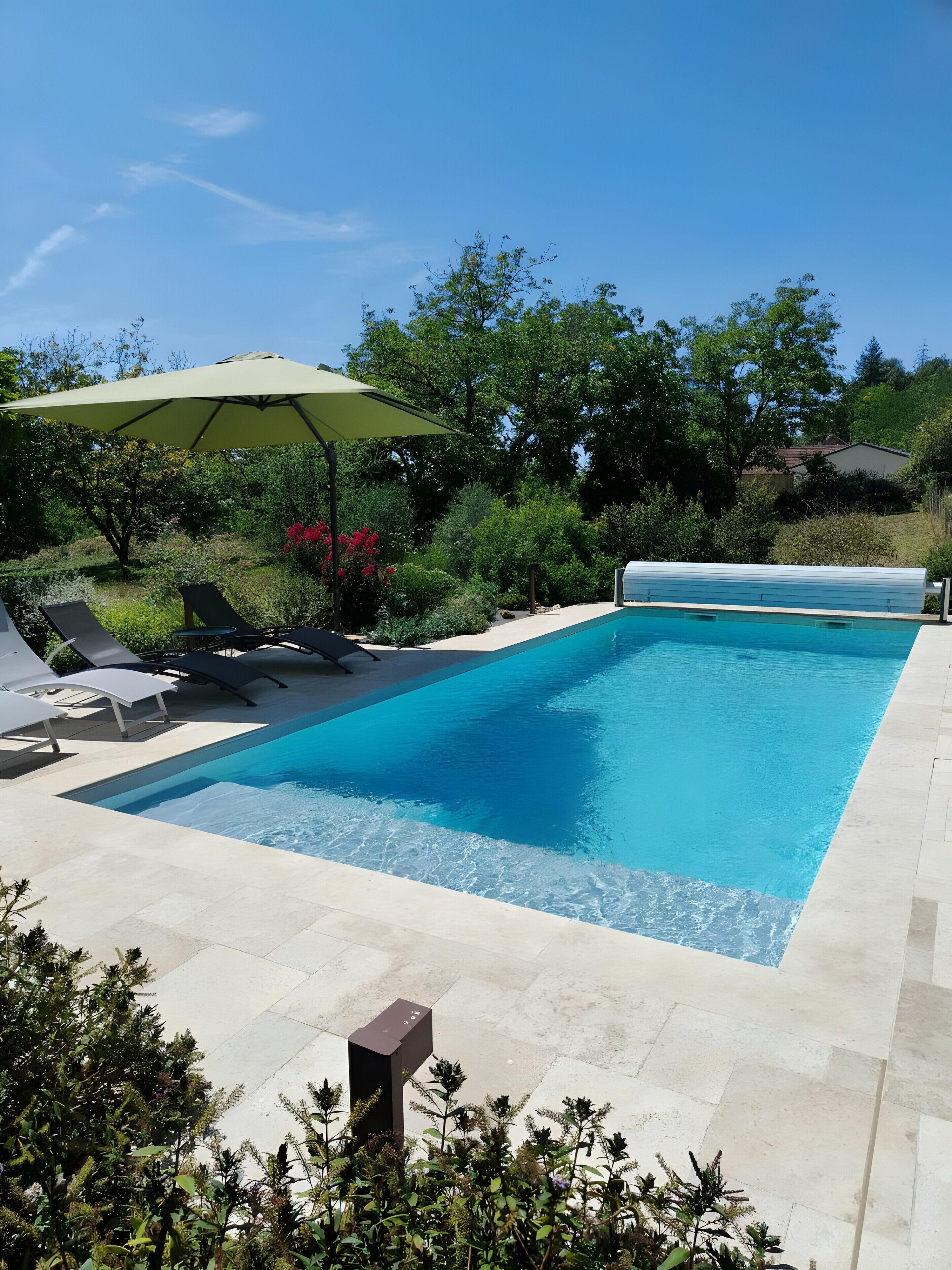 Ein rechteckiger Swimmingpool mit gefliestem Sonnendeck, umgeben von Grünpflanzen und einigen Liegestühlen.