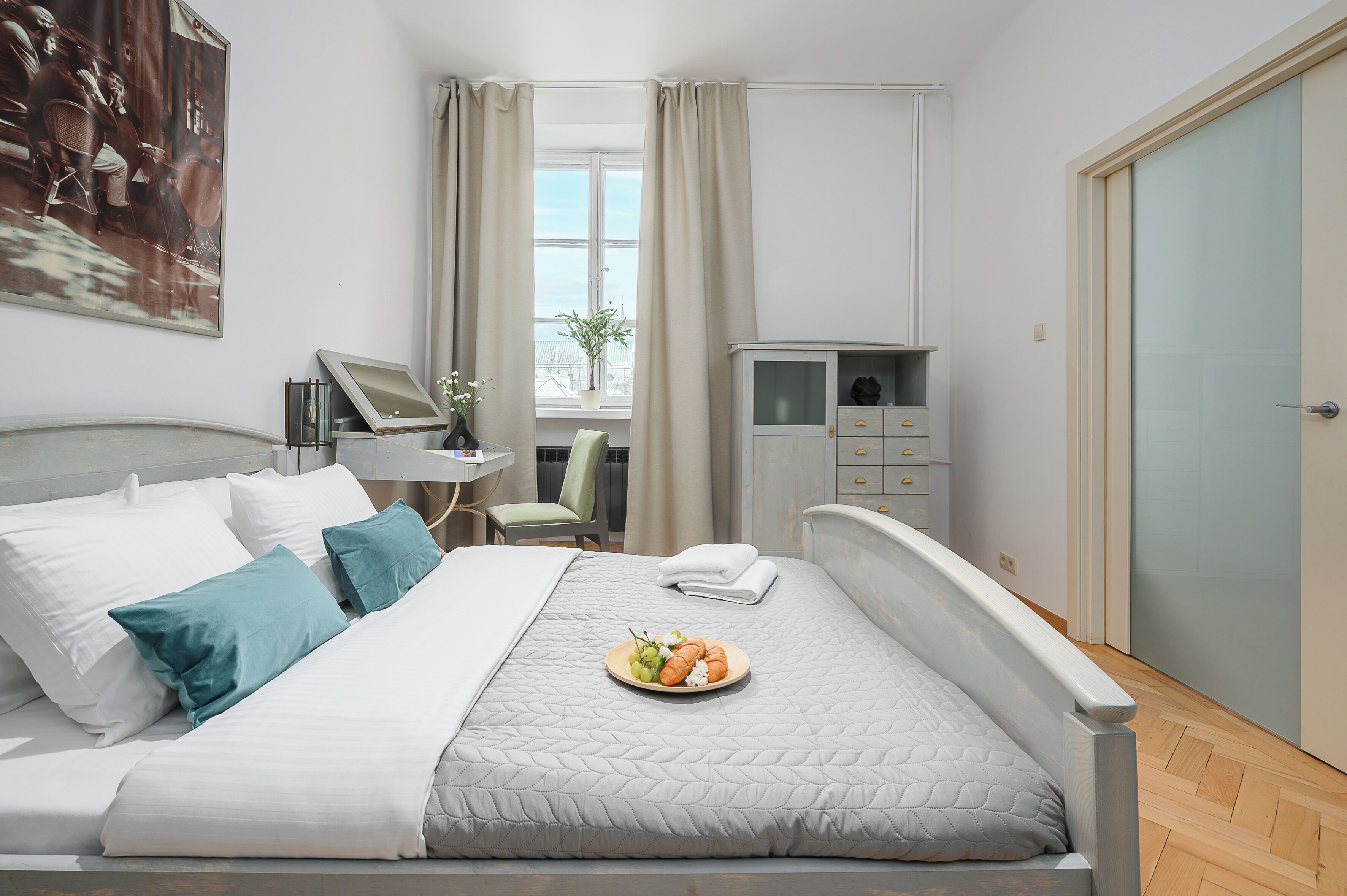 Una cama bien hecha con una bandeja de fruta, un escritorio con una silla, un armario y una ventana con cortinas.