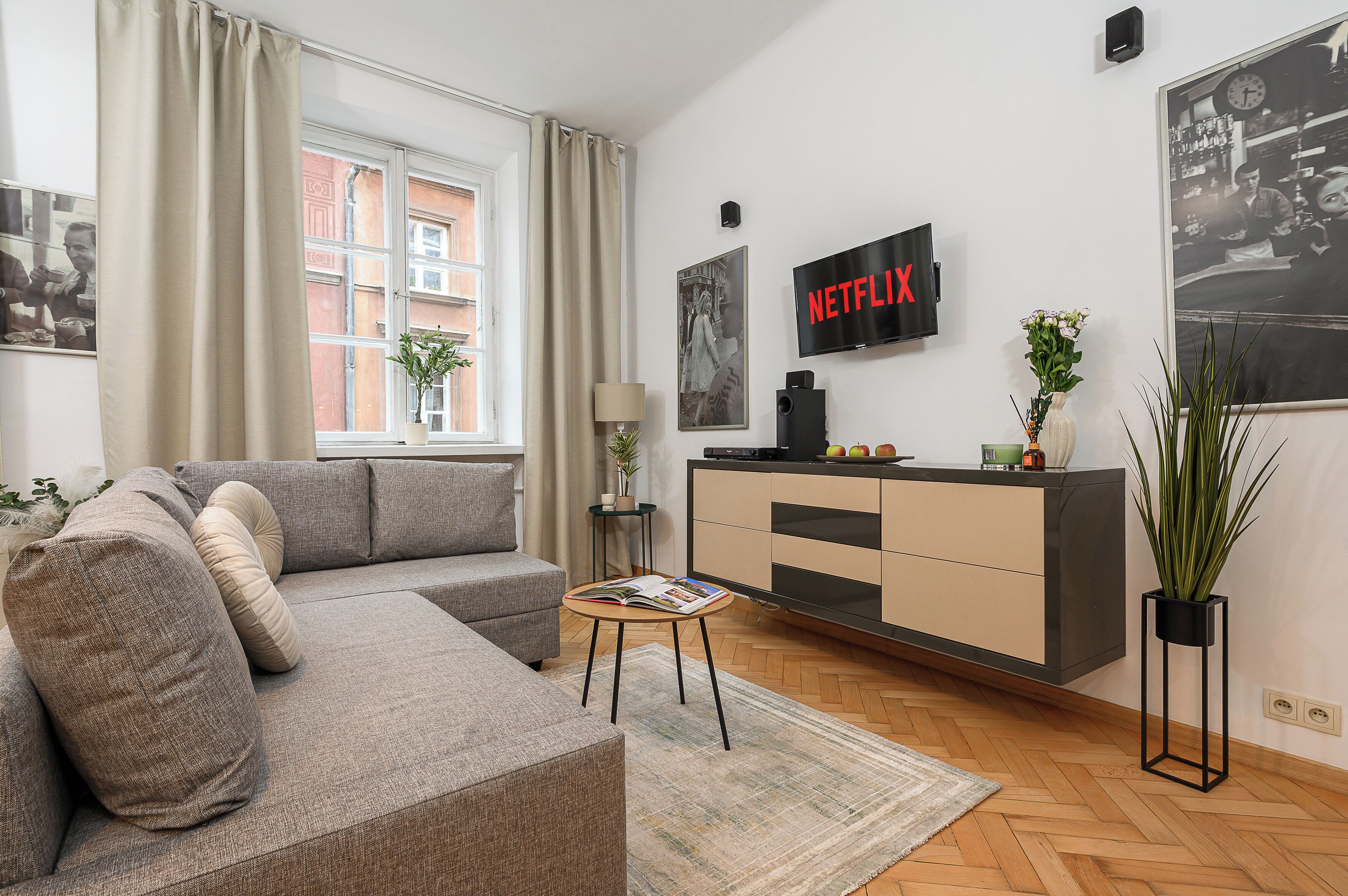 A modern living room with a grey sofa, a coffee table with a magazine, a TV mounted on the wall, and a potted plant.