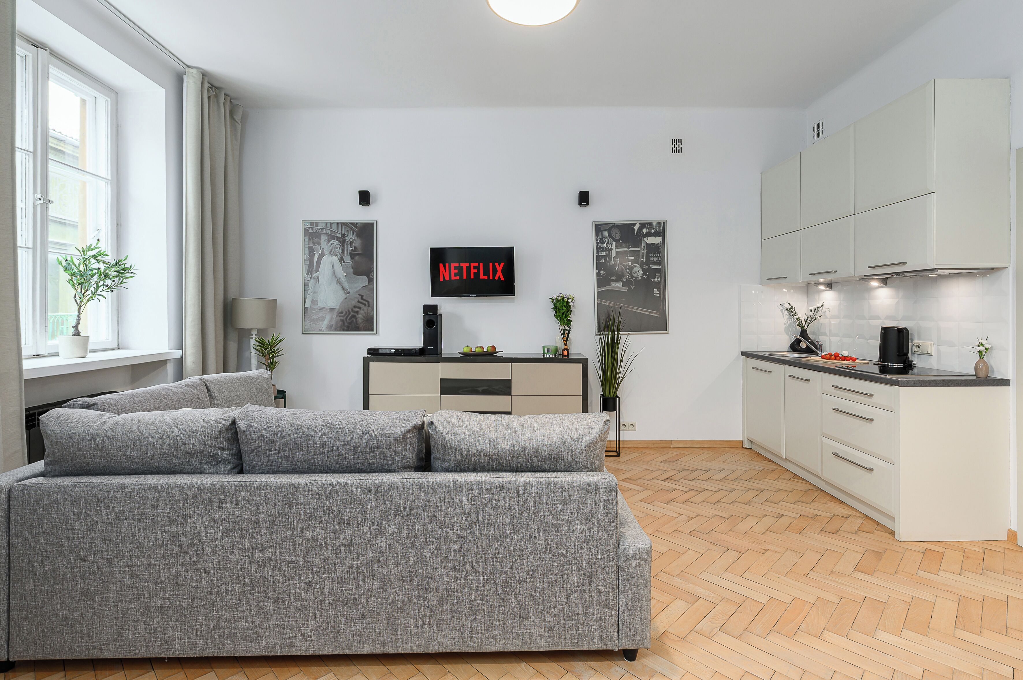 A modern living room with a grey sofa, a flat-screen TV, and a kitchen area with white cabinets.