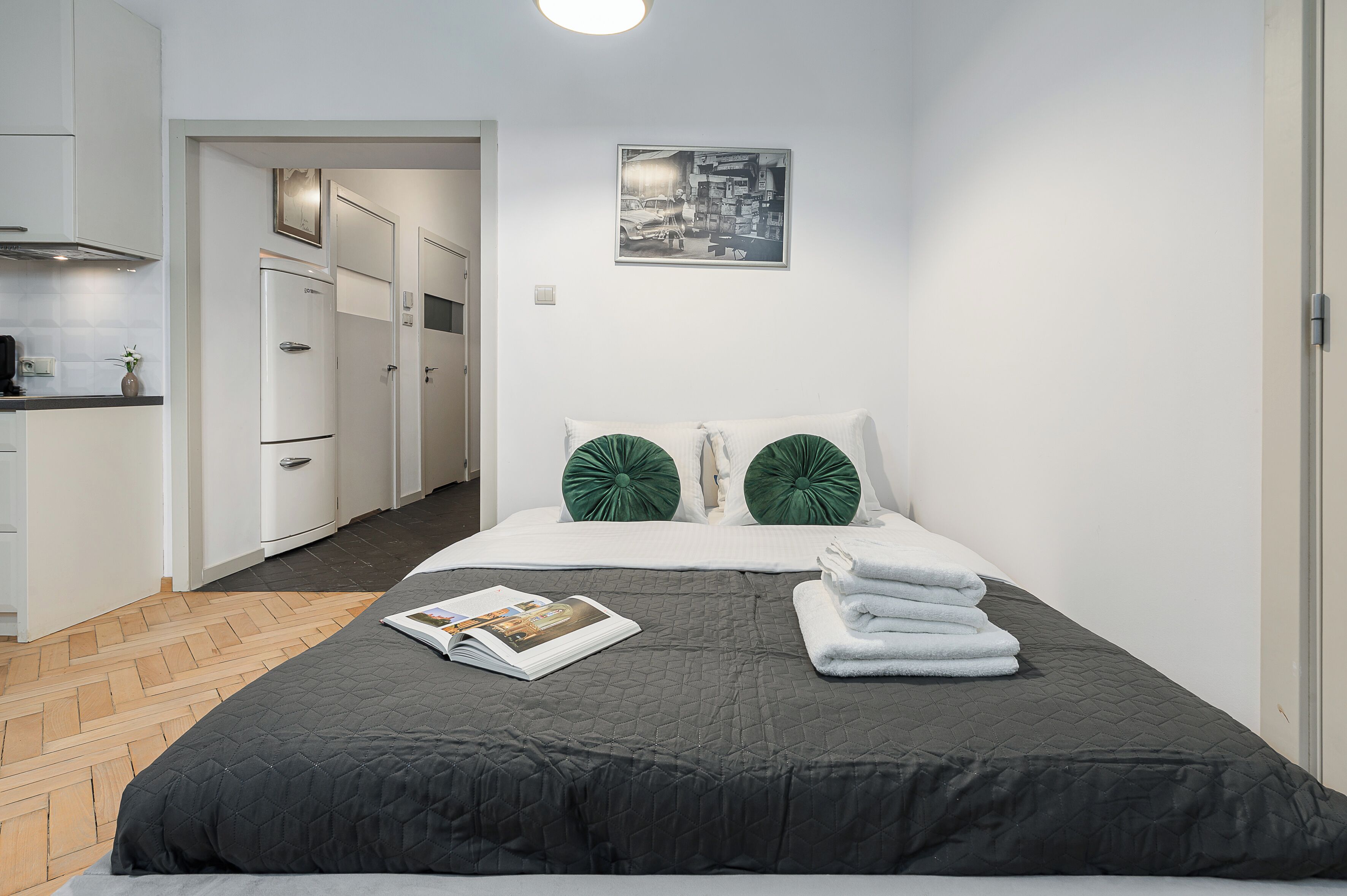 Una habitación de hotel con cama, una revista, toallas, un cuadro en la pared y una kitchenette al fondo.