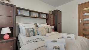 Une chambre d’hôtel avec un grand lit, une table de chevet avec une lampe, une commode, une étagère avec des livres et une plante, et une porte avec une fenêtre.