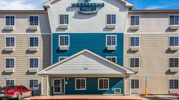 Ett hotell med flera våningar, ett tydligt skylt med texten ”Woodspring Suites”, en central entré och en parkeringsplats med en röd bil.