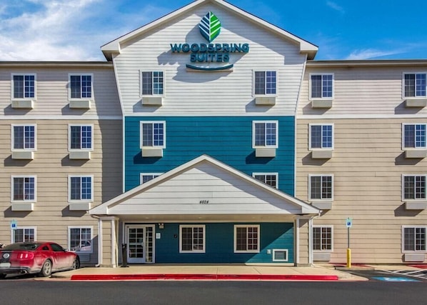 A multi-story hotel with a prominent 'Woodspring Suites' sign, a central entrance, and a parking lot with a red car. - Woodspring Suites Houston Baytown (Baytown)