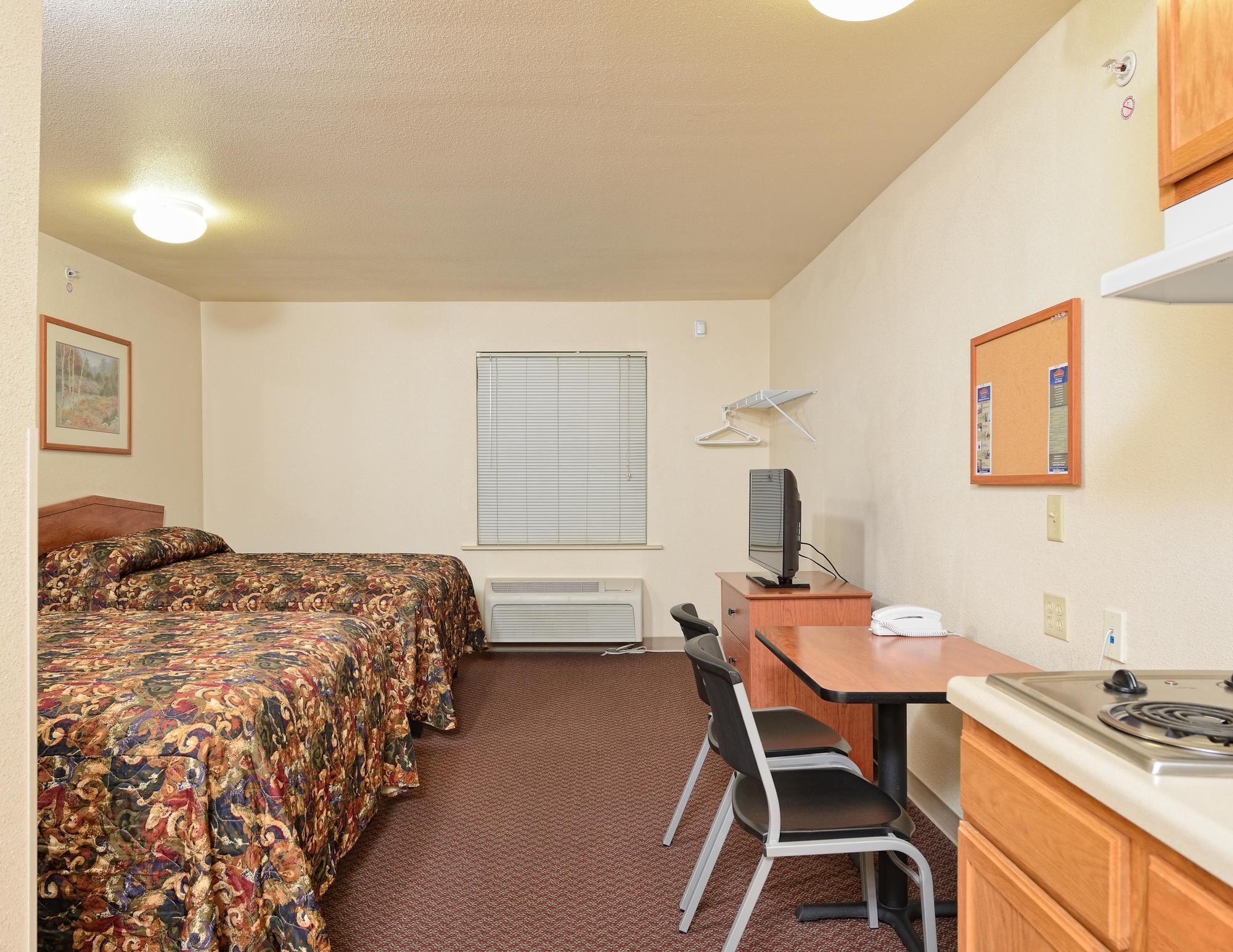 A small, well-lit room with two beds, a desk with a computer, a chair, a small table, and a stove.