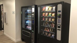 Deux distributeurs automatiques se trouvent dans un couloir, le long d’un mur blanc, et une porte est située sur le côté gauche.