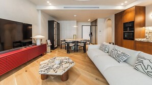 Uma sala de estar moderna com televisão de tela plana, um rack vermelho para entretenimento, uma mesa de centro de mármore, um sofá branco com almofadas estampadas e um espaço de jantar com mesa e cadeiras.