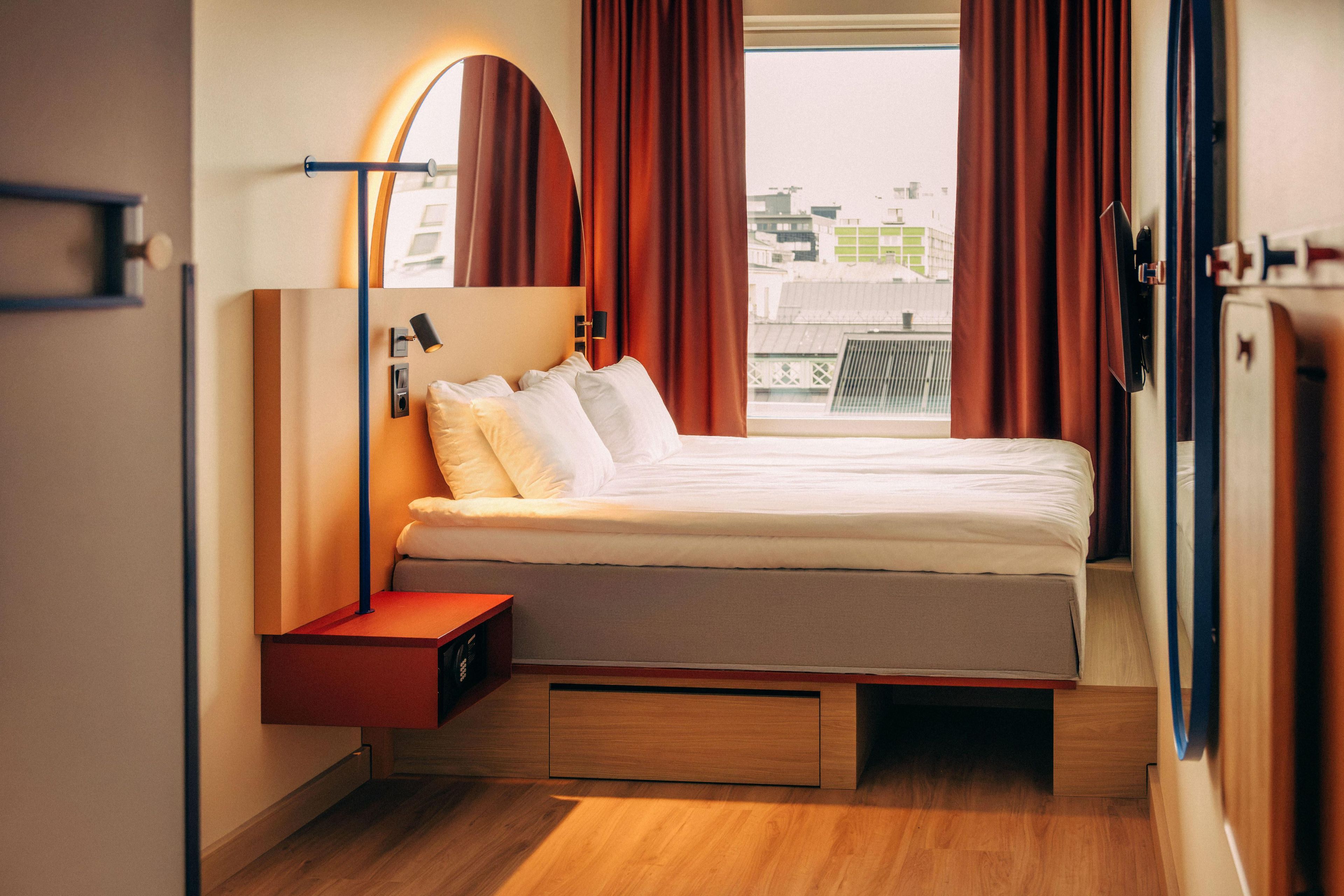 A modern hotel room with a bed, a side table, a mirror, and a view of the city.