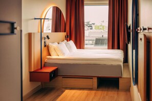 A modern hotel room with a bed, a side table, a mirror, and a view of the city.