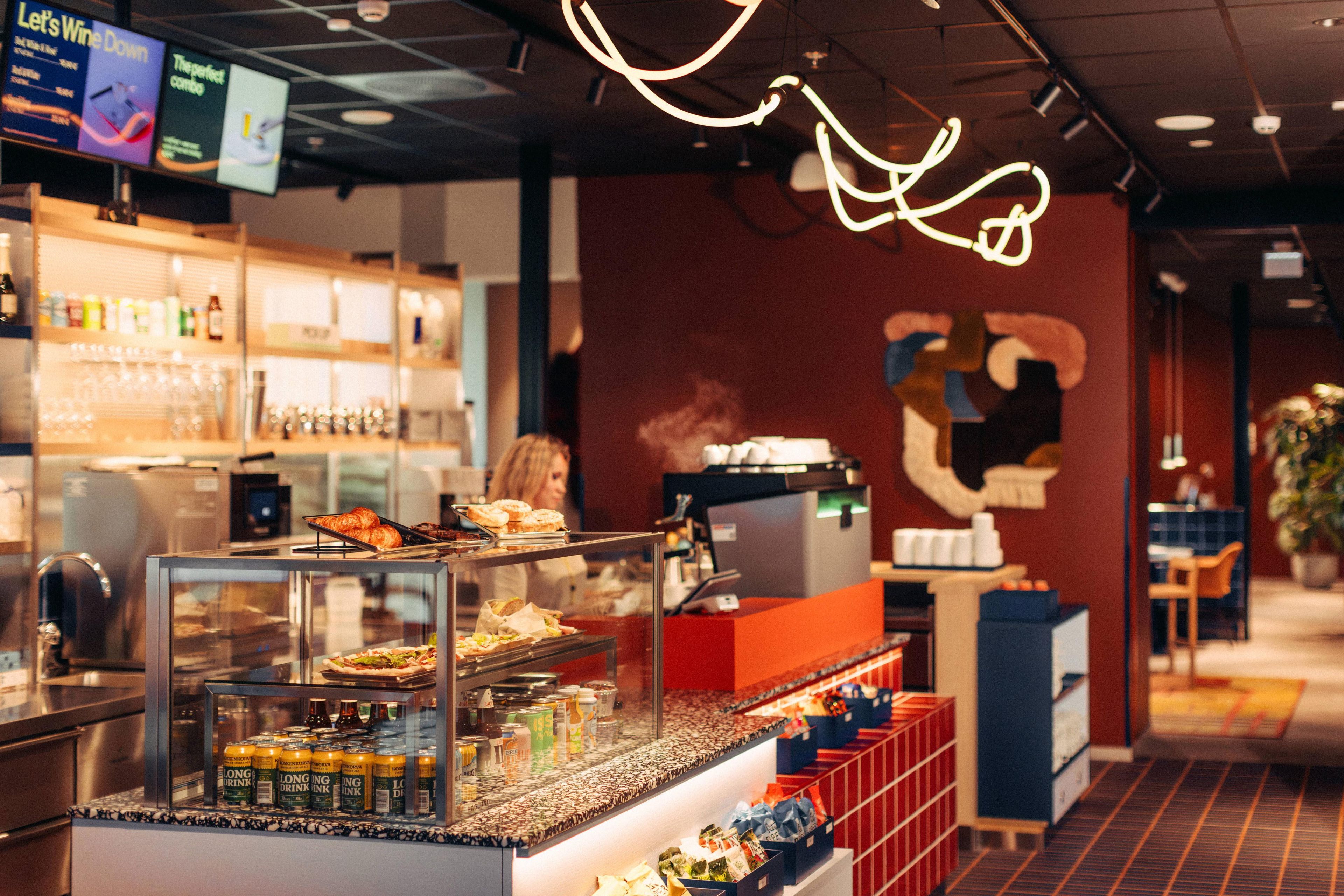A bar area with a counter displaying food and drinks, a menu board above, and a wall with a large abstract design.
