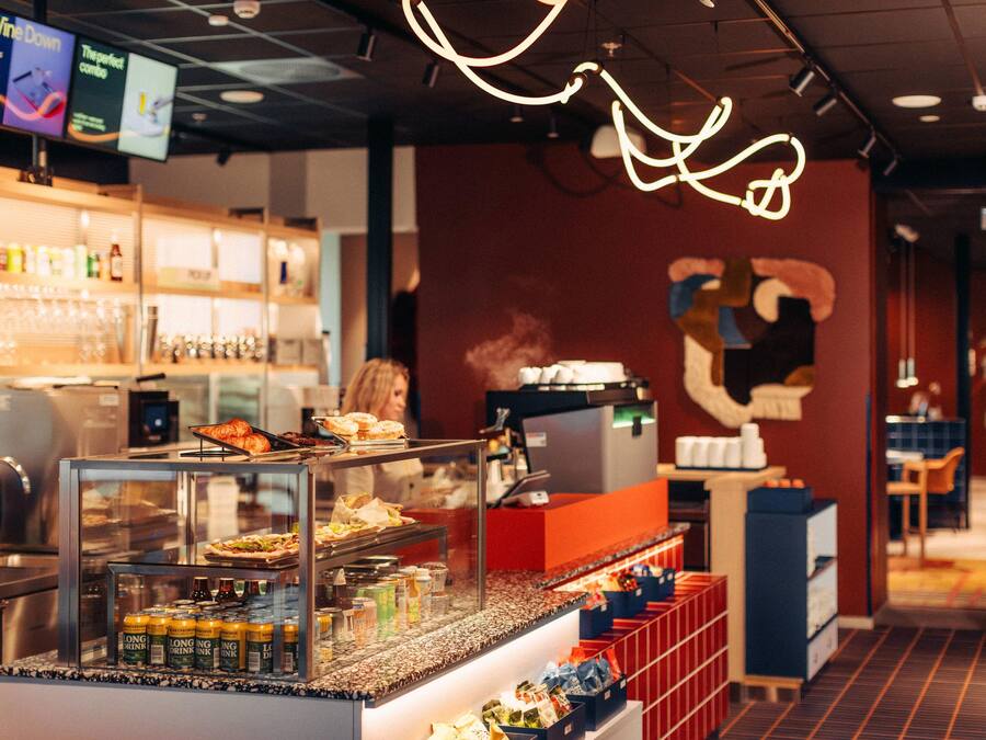 A bar area with a counter displaying food and drinks, a menu board above, and a wall with a large abstract design.