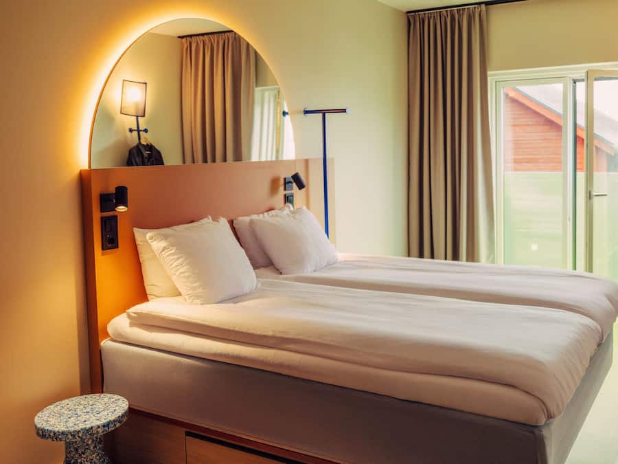 A double bed with a headboard, a small round table, and a chair in a hotel room with a view of the outdoors.