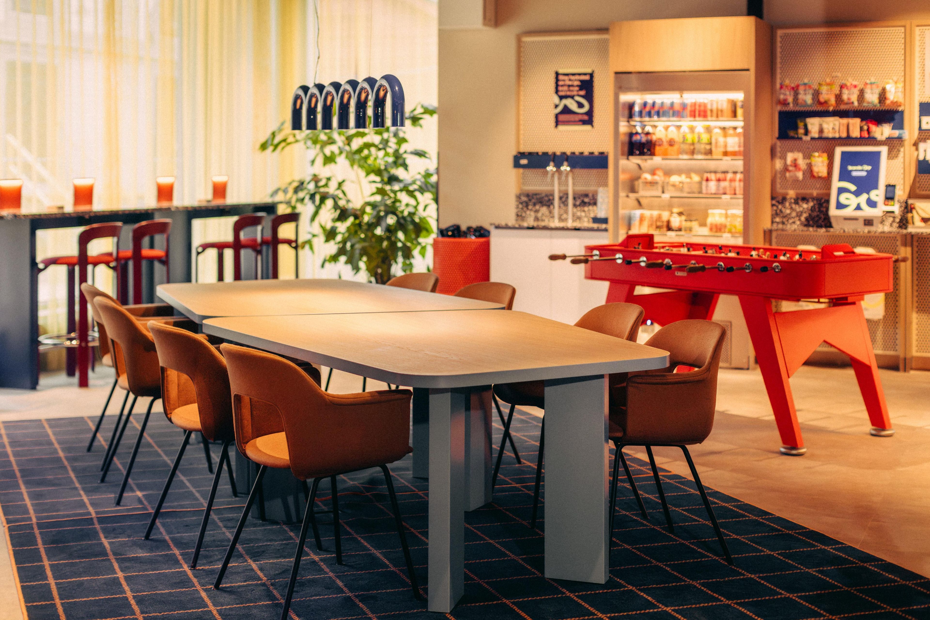 A restaurant with a long table, chairs, a foosball table, and a bar area with bottles.