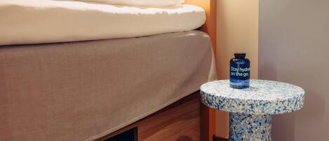A bed with a white comforter, a small blue table with a bottle, and a drawer with a black item.
