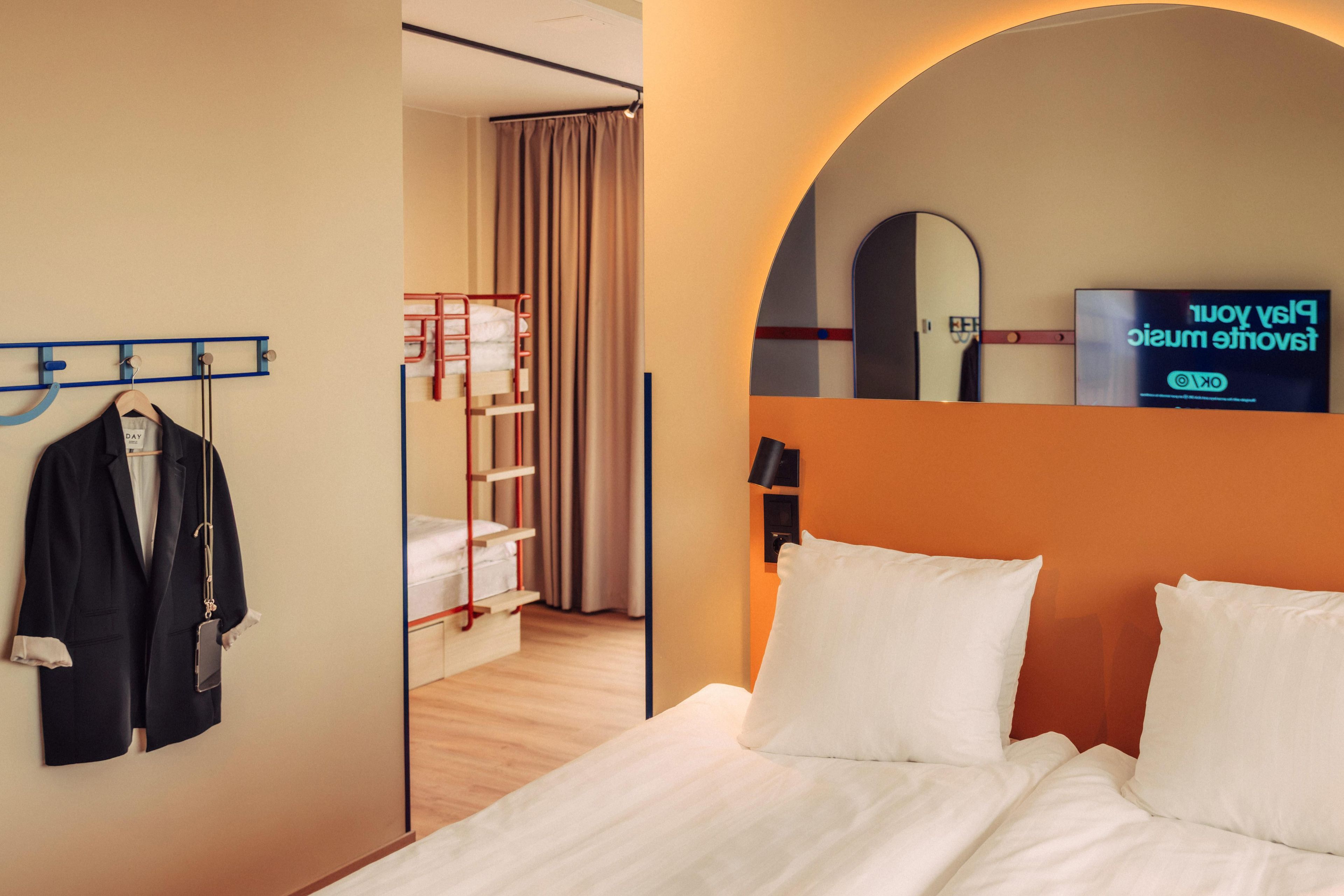 A hotel room with a bunk bed, a bed with white linens, a coat rack with a jacket, a mirror, and a sign on the wall.
