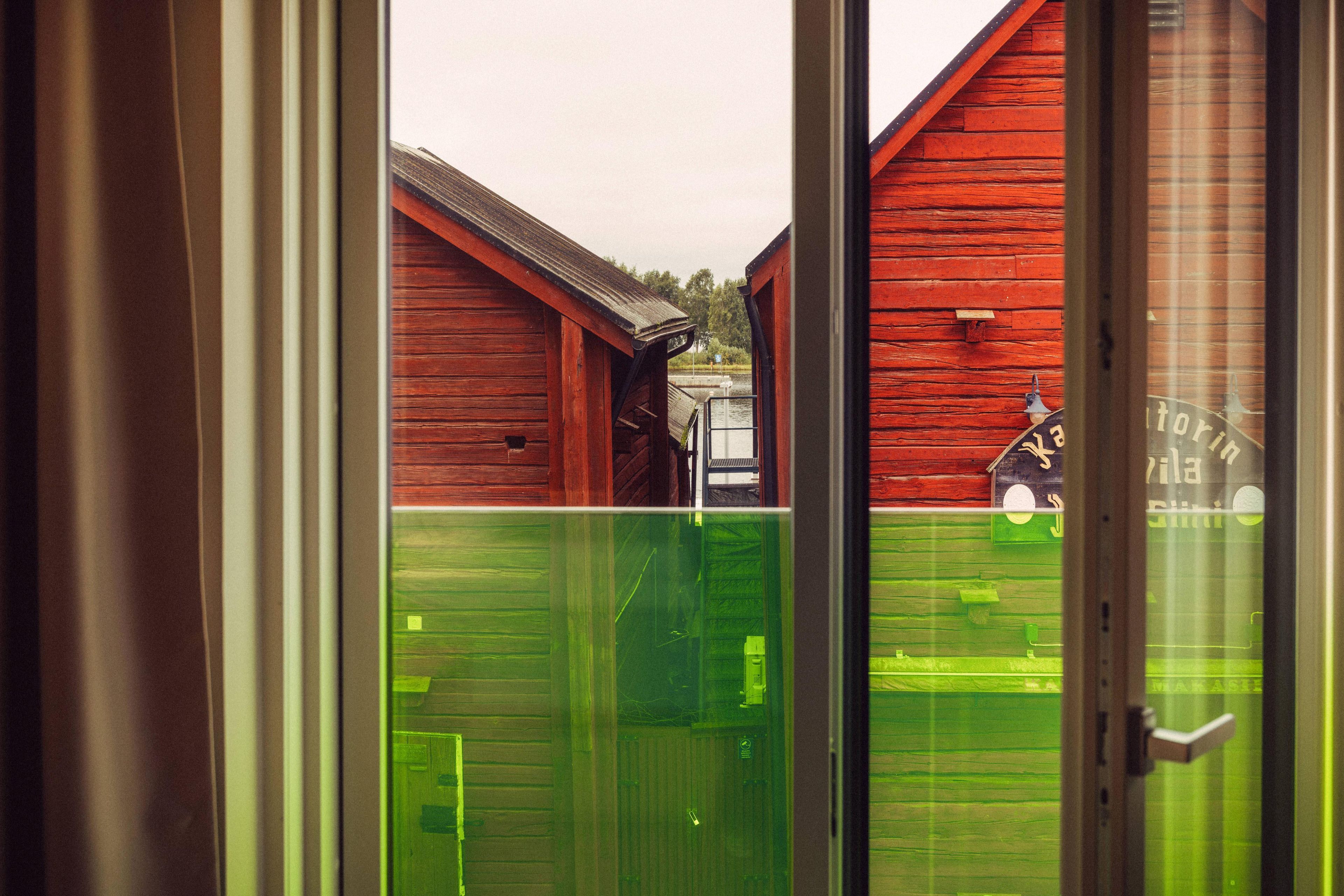On aperçoit, à travers une porte vitrée, un bâtiment en bois rouge avec un toit en métal, une clôture verte et une petite construction sur laquelle est apposée une enseigne.