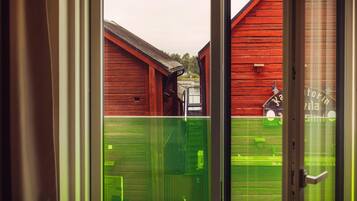 On aperçoit, à travers une porte vitrée, un bâtiment en bois rouge avec un toit en métal, une clôture verte et une petite construction sur laquelle est apposée une enseigne.