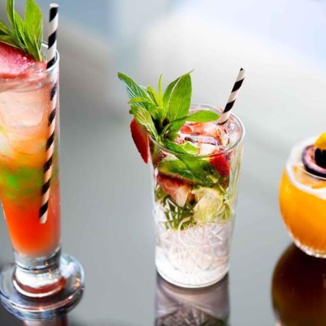 Two cocktails with straws and garnishes, one with mint leaves and the other with a slice of orange.