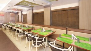 A long dining area with tables and chairs, a ceiling fan, and large windows with blinds. - Prajwal by Mango Hotels (Bengaluru)